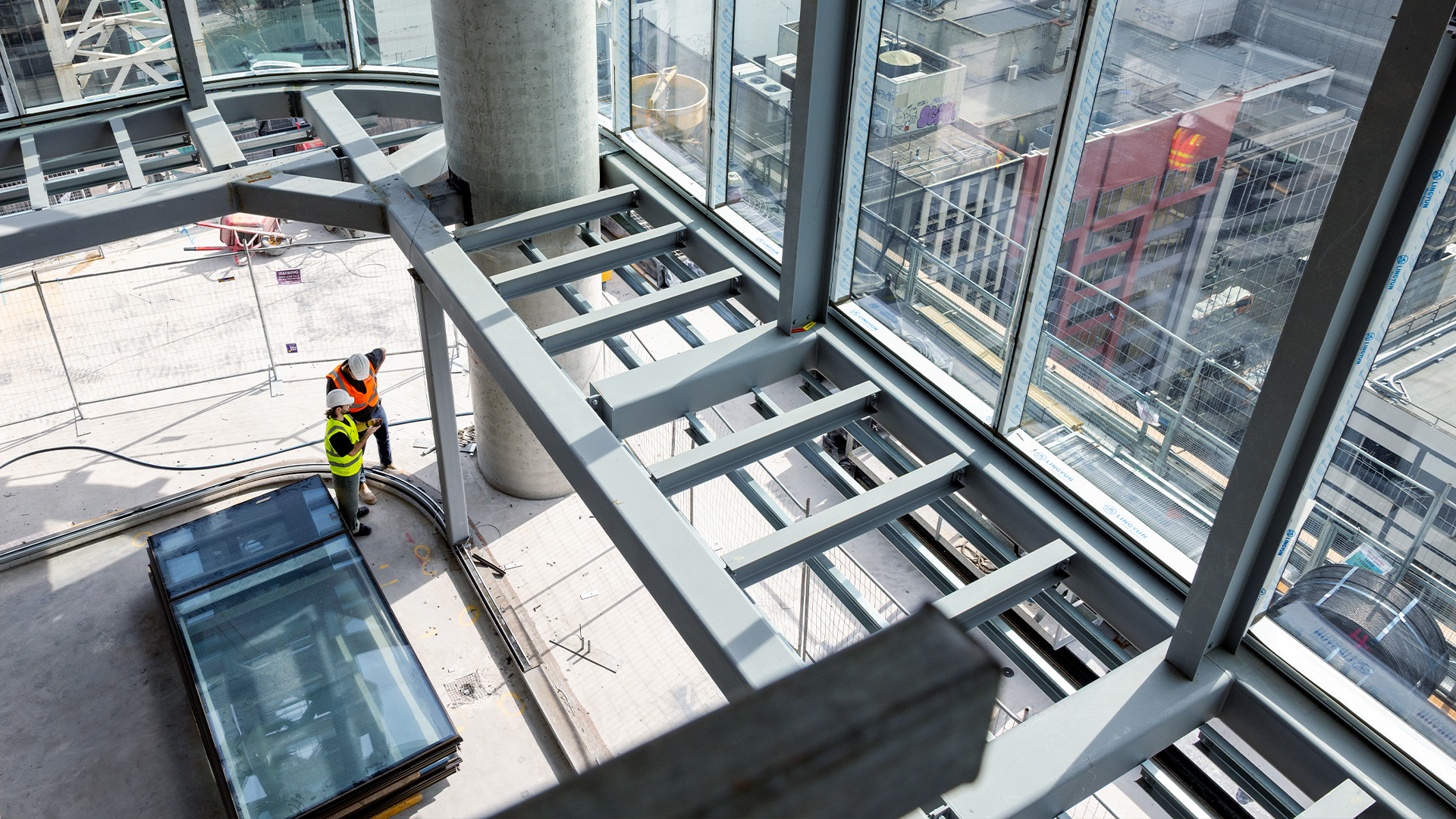 An elevated construction site interior is photographed from above, showing a large-scale development in an advanced stage of completion with structural steel framing, floor-to-ceiling glazing, and an expansive view of the surrounding built environment beyond the building envelope. A single worker in high-visibility clothing is visible on the floor below, illustrating the scale of the development and the complexity of managing post-completion defect management obligations across a building of this size. The image appears on Visibuild's post-completion software page to convey the scale and ambition of the construction projects that rely on structured defect liability period management systems to close out warranty items efficiently after handover. Large residential and commercial developments like the one pictured generate significant volumes of warranty defect tracking activity during the DLP period, making purpose-built DLP software construction tools essential for maintaining accountability across multiple trades, levels, and tenancies. Without a centralised construction handover software platform, managing the post-handover defect management workload across a development of this complexity would require a volume of manual coordination that is neither scalable nor auditable. This image reinforces the case for Visibuild's DLP management approach, positioning post-completion defect management software as a necessity for any contractor delivering large-scale construction projects with ongoing warranty management construction obligations.