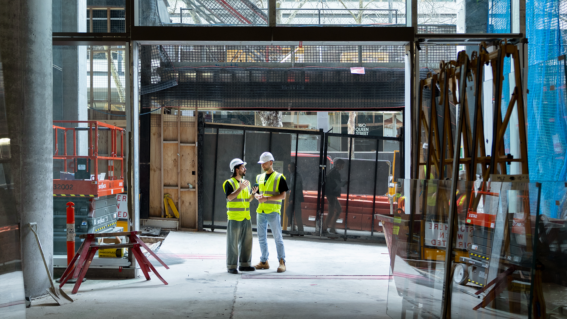 Two construction workers in high-visibility vests and white hard hats stand in conversation on an active construction site, surrounded by structural steel frames, large glazed door openings, scaffolding, and stacked building materials. The scene captures the kind of on-site coordination that underpins effective post-completion defect management — trade supervisors and project managers discussing remediation work, assigning tasks, and verifying progress during the defect liability period. The image is used on Visibuild's post-completion software page to represent the human and operational complexity of managing warranty defect tracking across a live construction environment where multiple trades are working simultaneously to resolve outstanding post-handover items. Without a structured DLP management platform connecting these on-site conversations to the office-based project team, critical warranty defect decisions risk being lost, undocumented, or miscommunicated. Visibuild's construction handover software bridges this gap by giving both site and office teams a shared, real-time view of all post-completion defect management activity — ensuring that every trade instruction, rectification update, and DLP tracking software interaction is captured and auditable. This image reinforces the core value of Visibuild's defect liability period management tools, illustrating that effective warranty management construction practice depends on clear communication between the people physically on site and the systems used to manage accountability from the office.