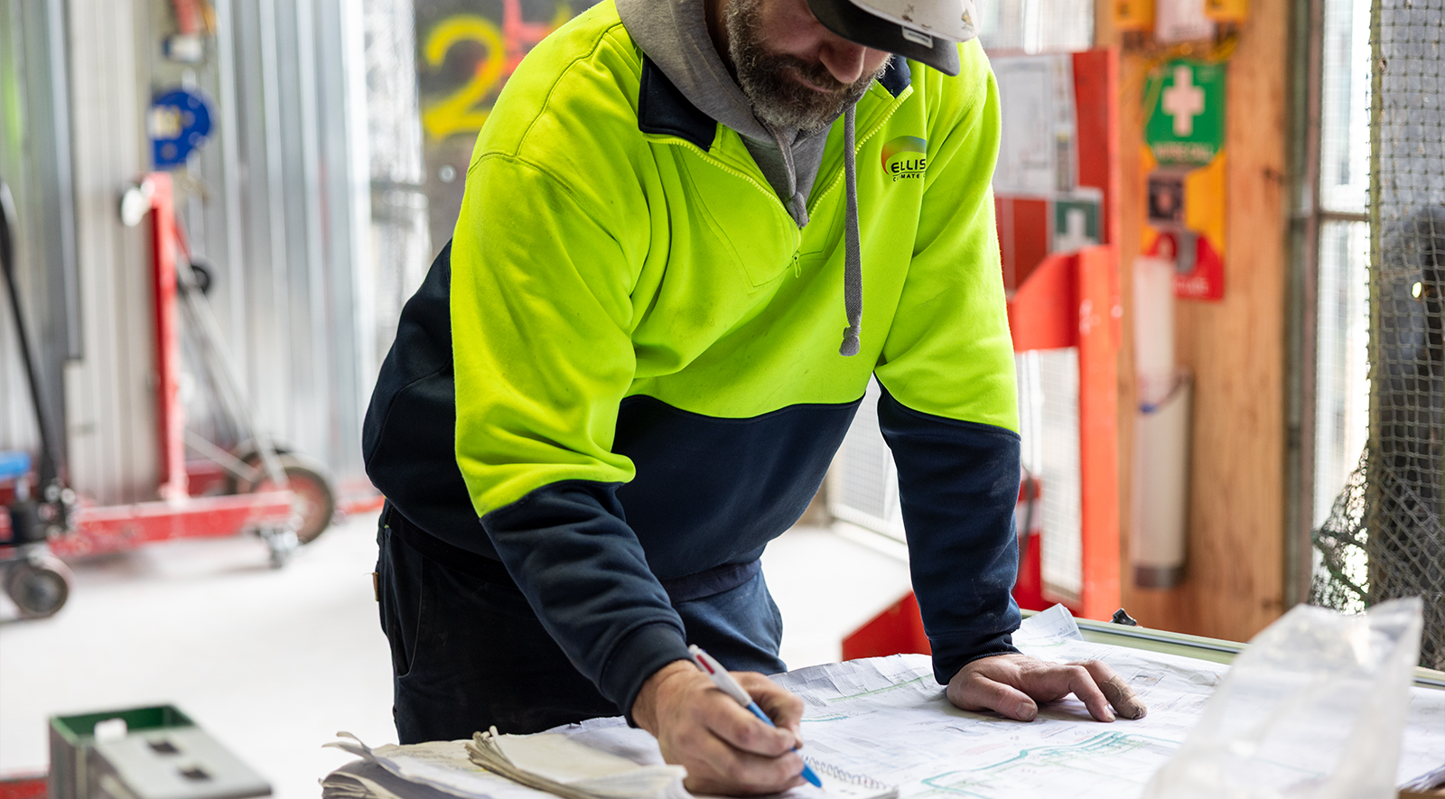 A subcontractor in a high-visibility yellow fleece jacket leans over a workbench on a construction site, reviewing printed plans or documentation with a pen in hand, surrounded by tools, safety equipment, and site materials in the background. The image is used on Visibuild's post-completion defect management page to represent the trade and subcontractor layer of the defect liability period management process — the on-site personnel who receive warranty defect assignments, carry out physical rectification work, and are accountable for supplying evidence of completion back to the head contractor. Effective DLP management depends on subcontractors receiving clear, timely instructions and responding with documented proof of resolution, yet this coordination is where post-handover defect management most commonly breaks down when managed through phone calls and email threads. Visibuild's DLP tracking software addresses this directly by enabling project managers to assign warranty items to specific trades from within the platform, set rectification requirements, and track response times — replacing the manual follow-up that consumes office team hours during the construction warranty software close-out process. This image reinforces the subcontractor accountability dimension of Visibuild's post-completion defect management platform, illustrating that structured warranty defect tracking benefits not just the office team but also the trades who need clear direction to complete DLP software construction obligations efficiently and on time.